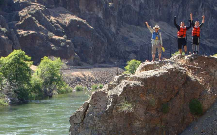 three people wearing life jackets stand on a rock above a river and raise their hands in celebration 